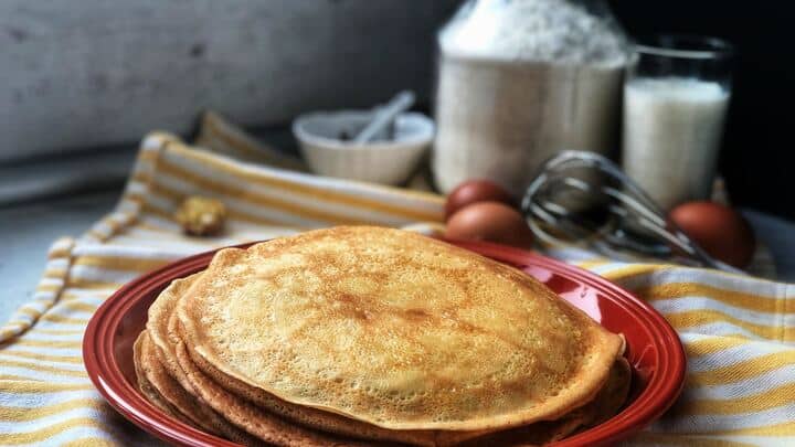 A stack of pancakes on a red plate. In the background, eggs, flour, milk, and a whisk are on a striped cloth.