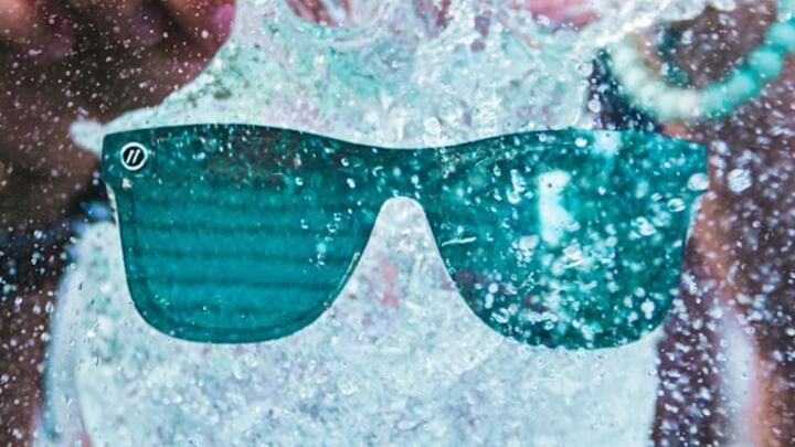 A pair of sunglasses is splashed with water, with droplets and motion visible on the lenses and in the background.