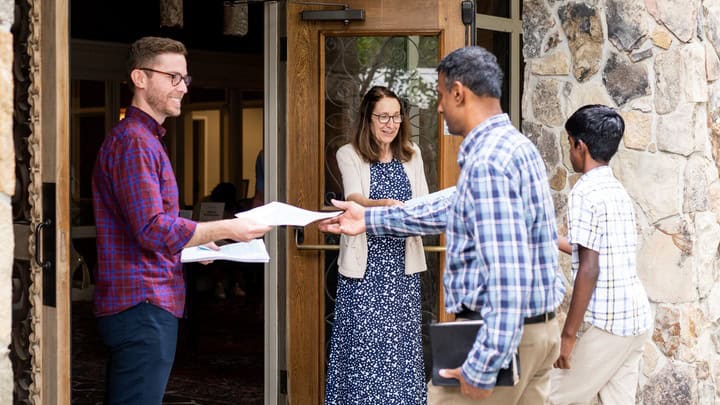 Two people stand at an open door handing out papers to individuals entering a stone building.